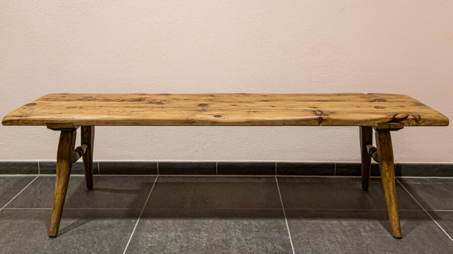 Bank aus Altholz ohne Lehne, mit natürlicher Baumkante und schrägen Beinen auf dunklem Steinboden.