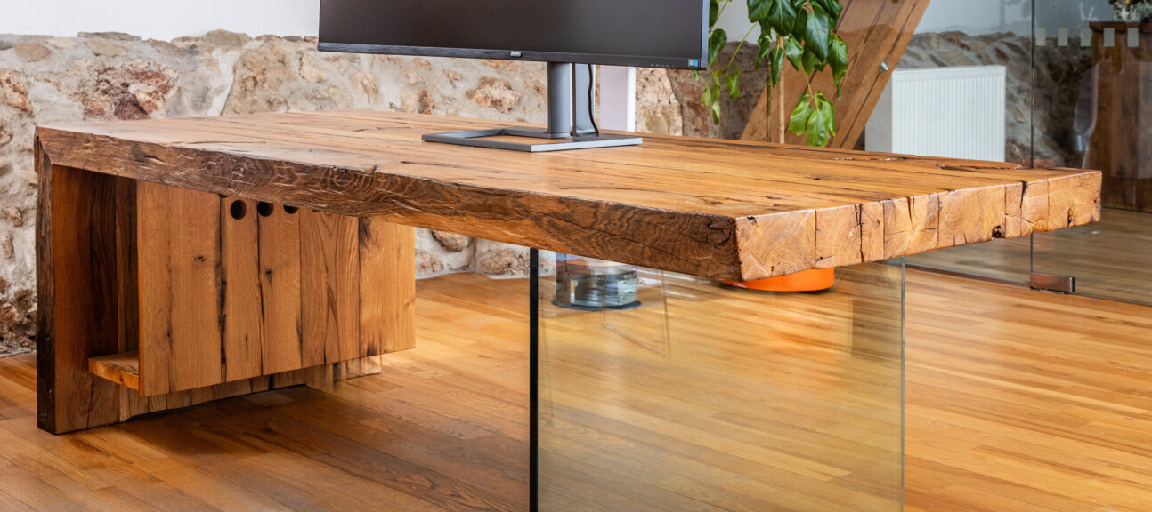 Schreibtisch aus Altholz mit Glasfuß und Curved-Monitor, in einem hellen Loftbüro.