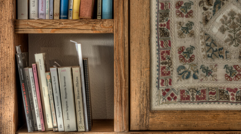 Ein Altholz-Bücherregal mit Büchern steht neben einem bestickten Tuch im Holzrahmen; das Arrangement schafft ein warmes, rustikales Interieur.