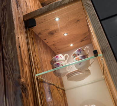 Beleuchtete Vitrine aus Altholz mit LED-Spots und Glasboden, darin gestapelte Teetassen und Untertassen.