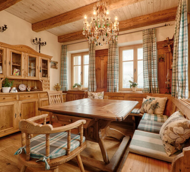 Eine gemütliche Bauernstube aus Altholz mit Eckbank, Tisch und Kredenz, holzgetäfelten Wänden, karierten Vorhängen und Kronleuchter.