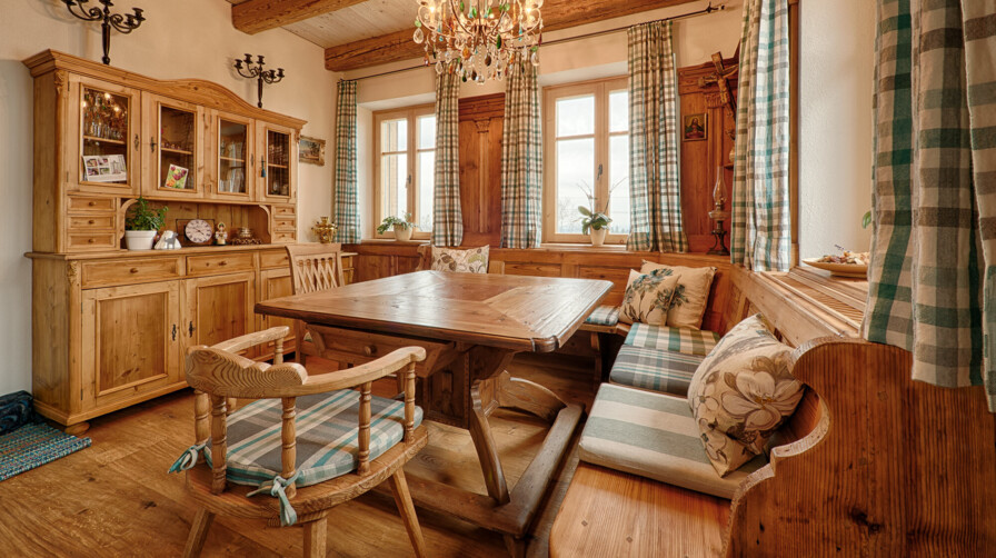 Eine gemütliche Bauernstube aus Altholz mit Eckbank, Tisch und Kredenz, holzgetäfelten Wänden, karierten Vorhängen und Kronleuchter.