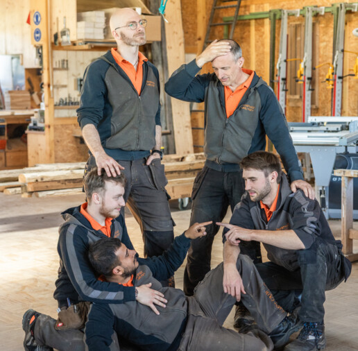 Teamfoto in der Antikholzwerkstatt: fünf Tischler posieren spielerisch in der Werkstatt zwischen Holzregalen, Werkzeugen und Maschinen.
