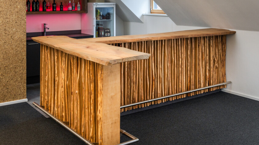 Bartheke aus Altholz, mit naturbelassener Kante und Stahl-Fußreling in modernem Raum; Kühlschrank und Flaschen im Hintergrund.