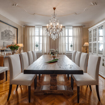 Elegantes Esszimmer mit Esstisch aus Altholz, weißen Polsterstühlen, Kristallkronleuchter und Glasvitrine auf Fischgrätparkett.