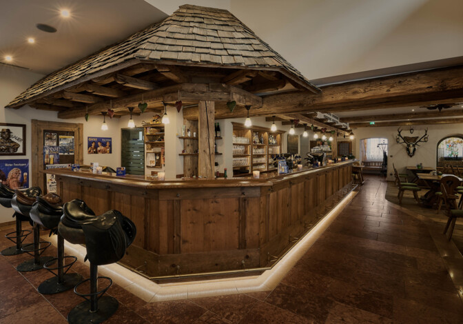 Uriges Barinterieur mit langer Holztheke, Balkendecke und Hockern; warm beleuchtete Möbel aus Altholz.