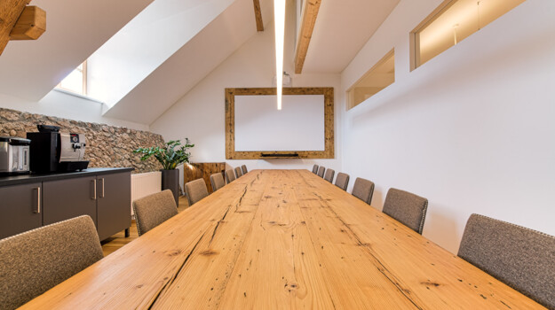 Besprechungsraum mit langem Konferenztisch aus Altholz, sowie Leinwand und Kaffeemaschine im Hintergrund.