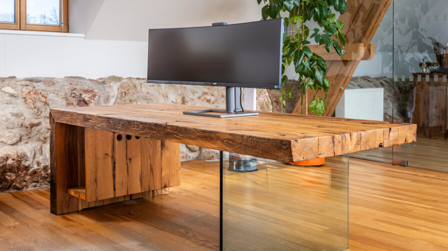 Schreibtisch aus Altholz mit Glasfuß und Curved-Monitor, in einem hellen Loftbüro.