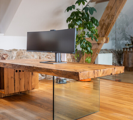 Schreibtisch aus Altholz mit Glasfuß und Curved-Monitor, in einem hellen Loftbüro.