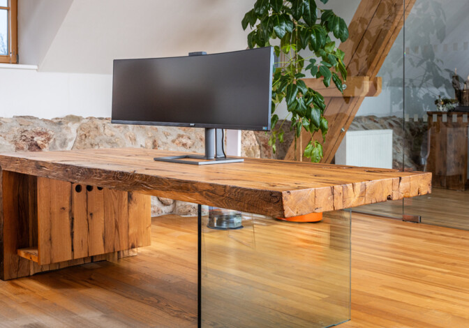 Schreibtisch aus Altholz mit Glasfuß und Curved-Monitor, in einem hellen Loftbüro.