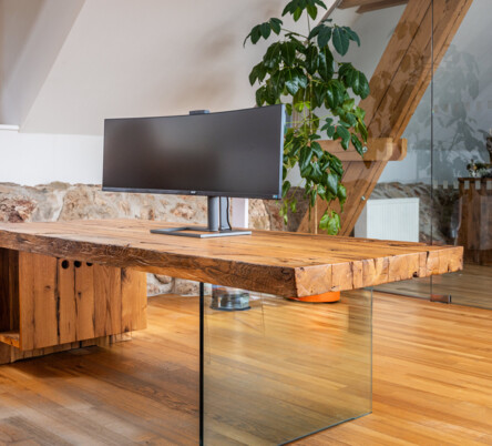 Schreibtisch aus Altholz mit Glasfuß und Curved-Monitor, in einem hellen Loftbüro.