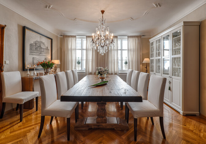 Elegantes Esszimmer mit Esstisch aus Altholz, weißen Polsterstühlen, Kristallkronleuchter und Glasvitrine auf Fischgrätparkett.