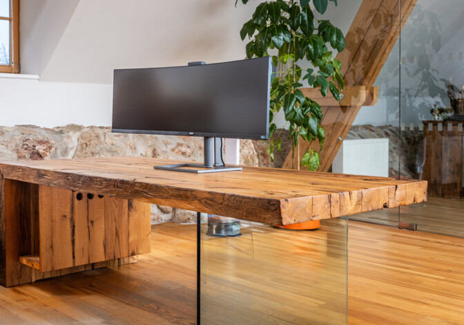 Schreibtisch aus Altholz mit Glasfuß und Curved-Monitor, in einem hellen Loftbüro.