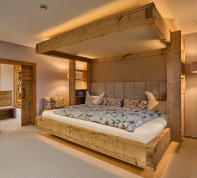 Gemütliches Schlafzimmer mit massivem Himmelbett aus Altholz, gepolstertem Kopfteil, integrierten Regalen und warmem Licht.