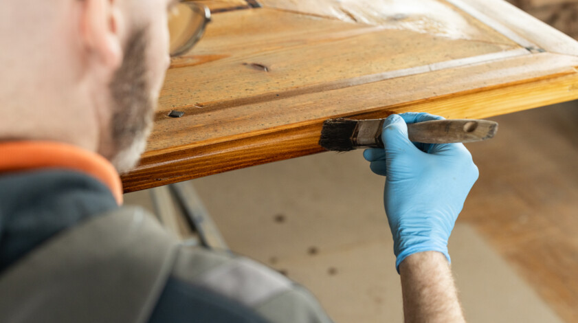 Handwerker trägt mit Pinsel Lack auf eine alte Holztür auf – sorgfältige Restaurierung antiker Holzoberflächen.