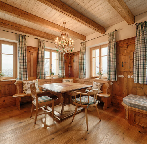 Gemütliche Bauernstube aus Altholz mit Sichtbalken, Eckbank, Holztisch, karierten Vorhängen und Kronleuchter.